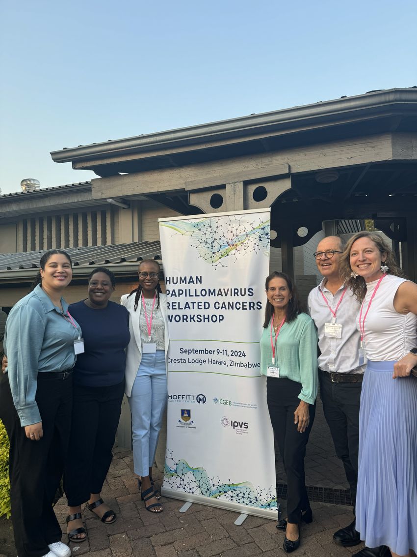 Six people stand surrounding a poster that says, “Human Papillomavirus Related Cancers Workshop.” Giuliano, on the immediate right of the poster, wears a green top and black pants.