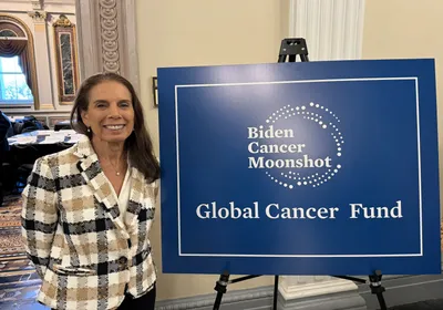 Anna Giuliano wears a checked blazer and stands next to a blue poster that says, “Biden Cancer Moonshot” and “Global Cancer Fund.”