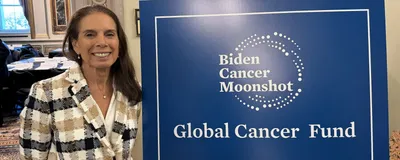 Anna Giuliano wears a checked blazer and stands next to a blue poster that says, “Biden Cancer Moonshot” and “Global Cancer Fund.”
