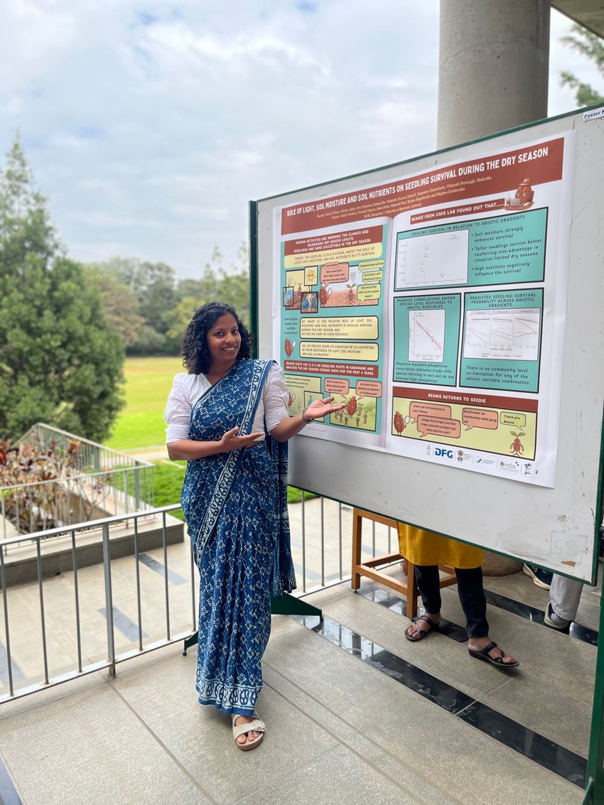Photograph of Vincy Wilson, a forest ecologist at the National Centre for Biological Sciences, standing in front of her comic-style poster at a science meeting at her institution. She is wearing a blue sari with a lighter colored pattern over a white blouse and has dark, shoulder-length hair. On her poster, there are panels similar to a comic book where the characters are having conversations as opposed to the information being contained in text blocks.
