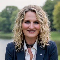Photograph of Carrie Wolinetz, who has blonde, shoulder length curly hair and is wearing a dark blazer over a white collared shirt. 