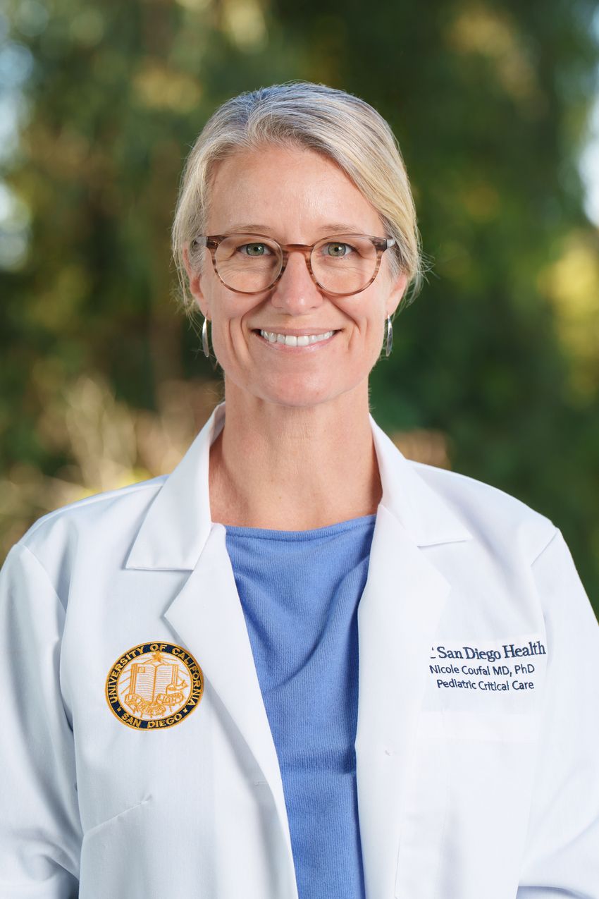 Photograph of Nicole Coufal, a physician scientist at the University of California, San Diego, standing in front of a blurred background of a tree. She is wearing a white lab coat with the University’s name, her name, her specialty, and the institution’s crest on it over a light blue top. She has light hair pulled back behind her head and is wearing glasses.
