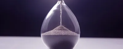 An hourglass stands upright with sand slowly flowing from the upper chamber to the lower one, indicating the accelerated aging in progeria.