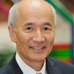 Photograph of Roger Wakimoto. He has grey and white hair on the sides of his head and is wearing a dark suit with a white collared shirt. 