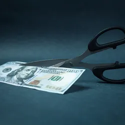 Photo of a pair of black-handled scissors set to cut a US one hundred dollar bill in half on a blue background. 