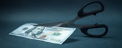 Photo of a pair of black-handled scissors set to cut a US one hundred dollar bill in half on a blue background. 