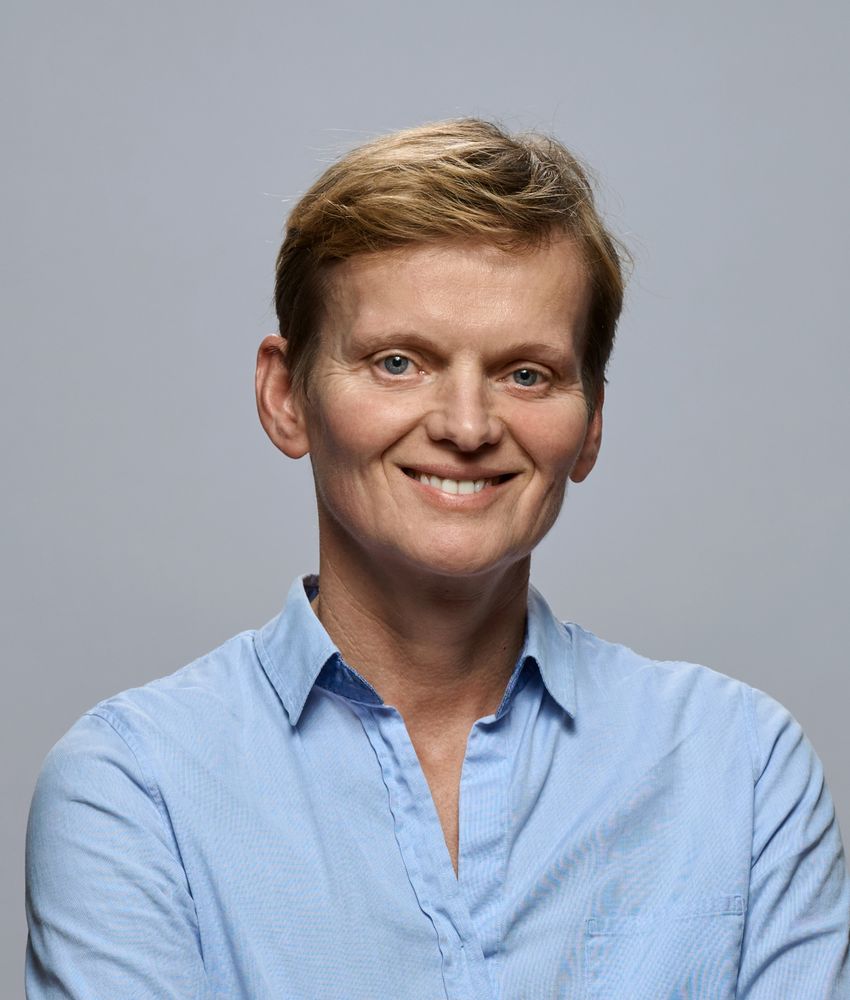 A photograph of Ruth Loos, a human geneticist at the University of Copenhagen, standing in front of a grey background. Loos has short, light hair and is wearing a blue collared shirt and is smiling at the camera.
