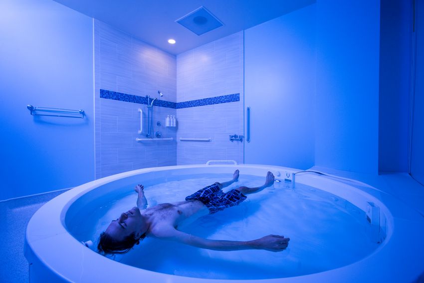 A person floats in a shallow floatation tank in a blue-lit room.