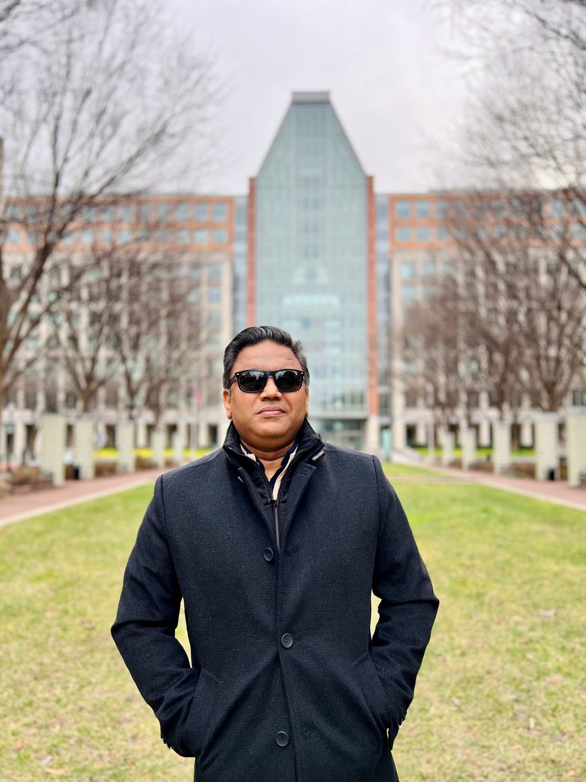 A photograph of Masudur Rahman, a patent examiner for the United States Patent and Trademark Office, standing in front of the USPTO building. Rahman has short dark hair and is wearing dark sunglasses. He has his hands in the pockets of a long, black coat that he is wearing.