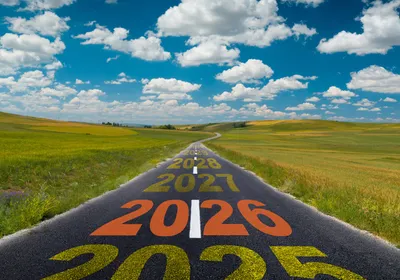 Image of a road cutting through a field under a blue sky with clouds. On the road, the years “2025”, “2026”, “2027”, “2028” are painted leading down the road. 2026 is in orange. This represents the outlook for research in 2026.