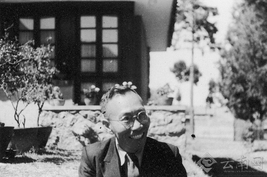 Tang Feifan smiles and wears glasses in a black and white photo taken in front of a house.