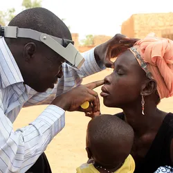 A technician performs an eye exam on a woman as she holds a child in a training exercise outside of Ouagadougou, Burkina Faso.
