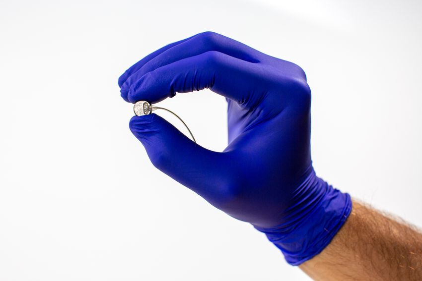 A person wears a blue glove while holding a piece of a brain-computer interface against a white background. A person wears a blue glove while holding a piece of a brain-computer interface against a white background.