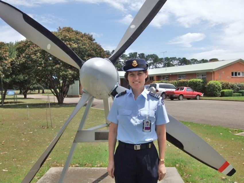 Kirsty Wright is wearing an air force uniform of light blue shirt, navy pants, and hat, with insignia for her rank of squadron leader on her shoulders. She stands outside, in front of an airplane propeller.