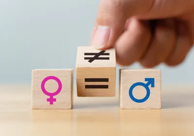 Wooden blocks with symbols of the male (blue, right) and female (pink, left) sex lay on a table. A person’s hand holds another block, which has an equal sign on one side and an inequal sign on another, in the middle.