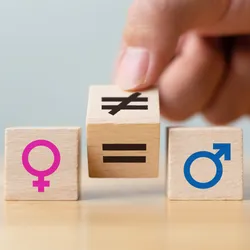 Wooden blocks with symbols of the male (blue, right) and female (pink, left) sex lay on a table. A person’s hand holds another block, which has an equal sign on one side and an inequal sign on another, in the middle.