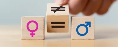 Wooden blocks with symbols of the male (blue, right) and female (pink, left) sex lay on a table. A person’s hand holds another block, which has an equal sign on one side and an inequal sign on another, in the middle.