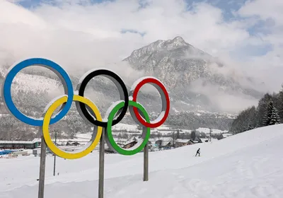 A statue of the Olympic Rings covered in a dusting of snow with a mountain and a cross-country ski track in the background.
