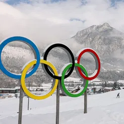 A statue of the Olympic Rings covered in a dusting of snow with a mountain and a cross-country ski track in the background.