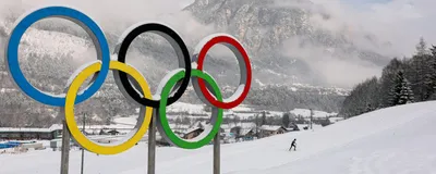 A statue of the Olympic Rings covered in a dusting of snow with a mountain and a cross-country ski track in the background.