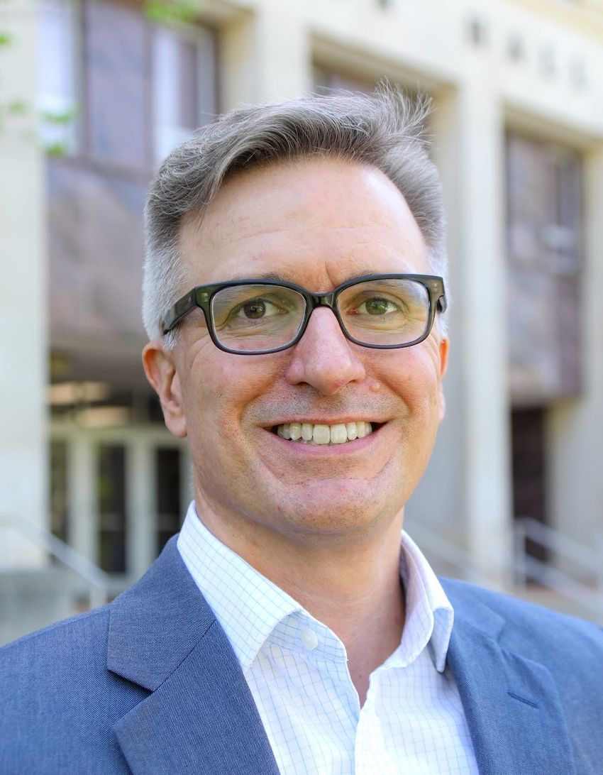 Photograph of Michael Vitevitch, a cognitive psychologist studying speech errors to understand language and hearing disorders. He is smiling at the camera and has short, light-colored hair. He is wearing black glasses and a blue suit jacket over a light collared shirt with a square pattern.