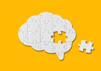 Photograph of a white puzzle in the shape of a brain on a yellow background. One puzzle piece from the center of the brain is missing. This photo represents a word losing its meaning in a phenomenon called semantic satiation. 