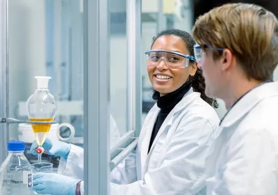 A scientist is working in a chemical fume hood as she looks over at her colleague. Both are wearing white lab coats in the laboratory.