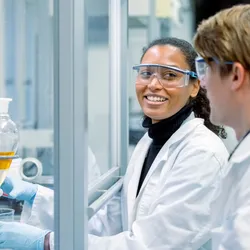 A scientist is working in a chemical fume hood as she looks over at her colleague. Both are wearing white lab coats in the laboratory.
