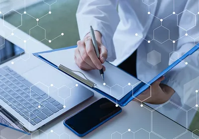 A photo of a scientist in a lab coat writing notes while in front of a laptop and cellphone