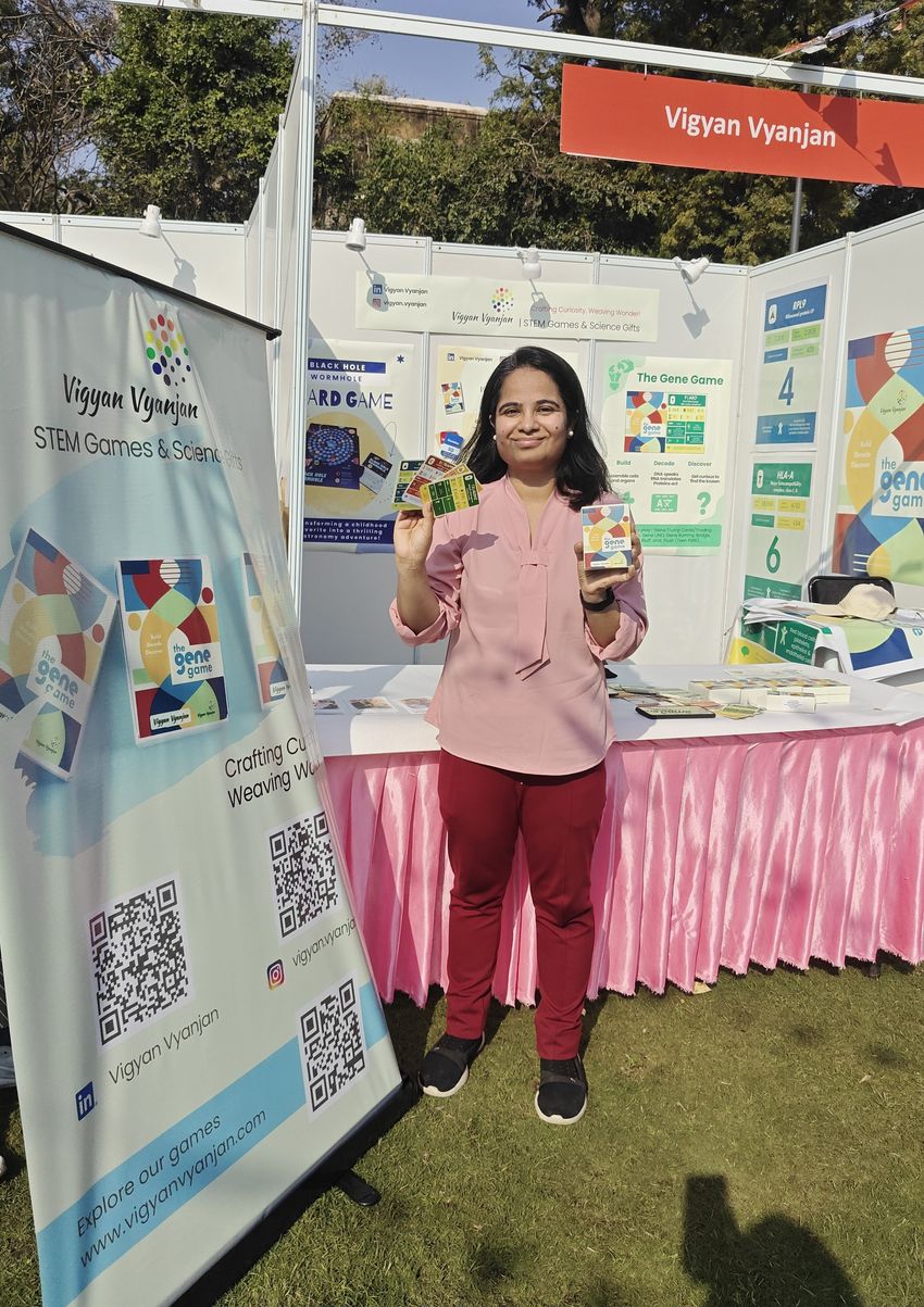 Poorti Kathpalia, wearing a pink top and red pants, holds The Gene Game in her hands in front of a booth labelled “Vigyan Vyanjan.” Poorti Kathpalia, wearing a pink top and red pants, holds The Gene Game in her hands in front of a booth labelled “Vigyan Vyanjan.”