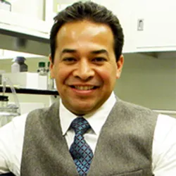 Ivan Martinez wears a grey vest over a white collared shirt with a blue tie in a research laboratory.