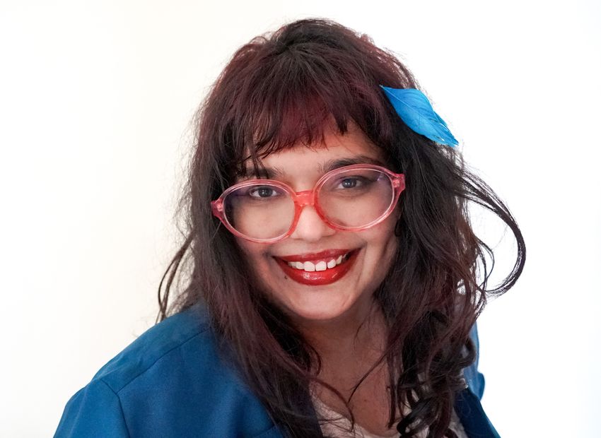 A photograph of Upasana Sarraju wearing a blue top and pink glasses. She wears a blue feather in her hair.