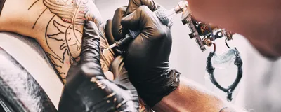 A tattoo artist, wearing black gloves, works on a client’s left arm.   
