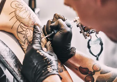 A tattoo artist, wearing black gloves, works on a client’s left arm.   