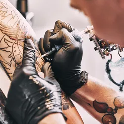 A tattoo artist, wearing black gloves, works on a client’s left arm.   