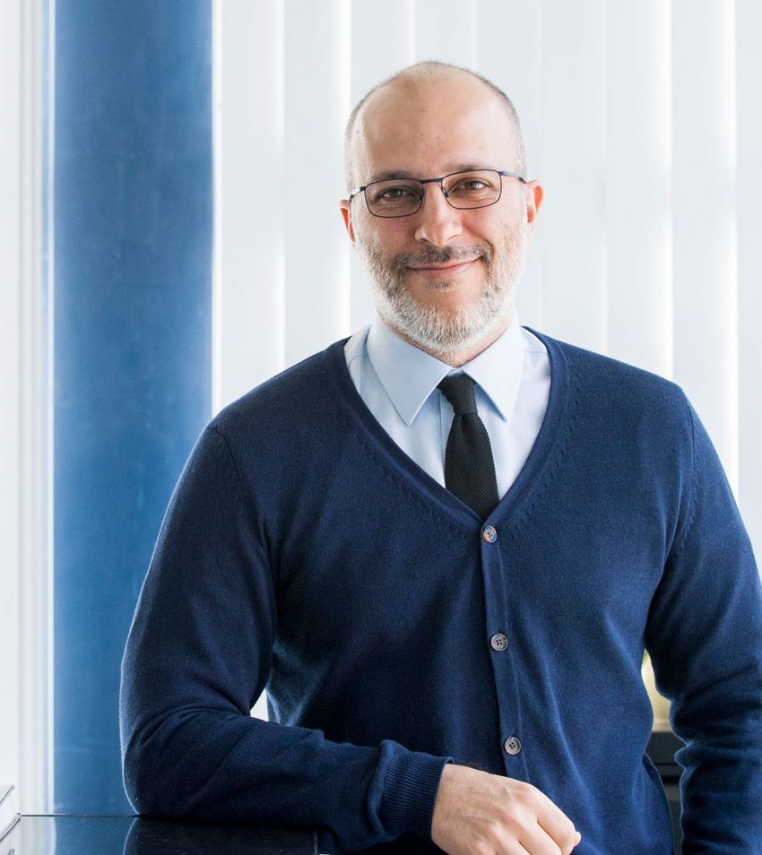 Santiago González, wearing a dark blue sweater, light blue shirt, black tie, and glasses, poses in front of a blue wall and white blinds.