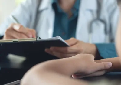 A young child sits with their arms crossed in the foreground as a doctor writes something on a clipboard.