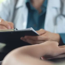 A young child sits with their arms crossed in the foreground as a doctor writes something on a clipboard.
