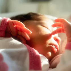 A newborn baby sleeps while touching her face.
