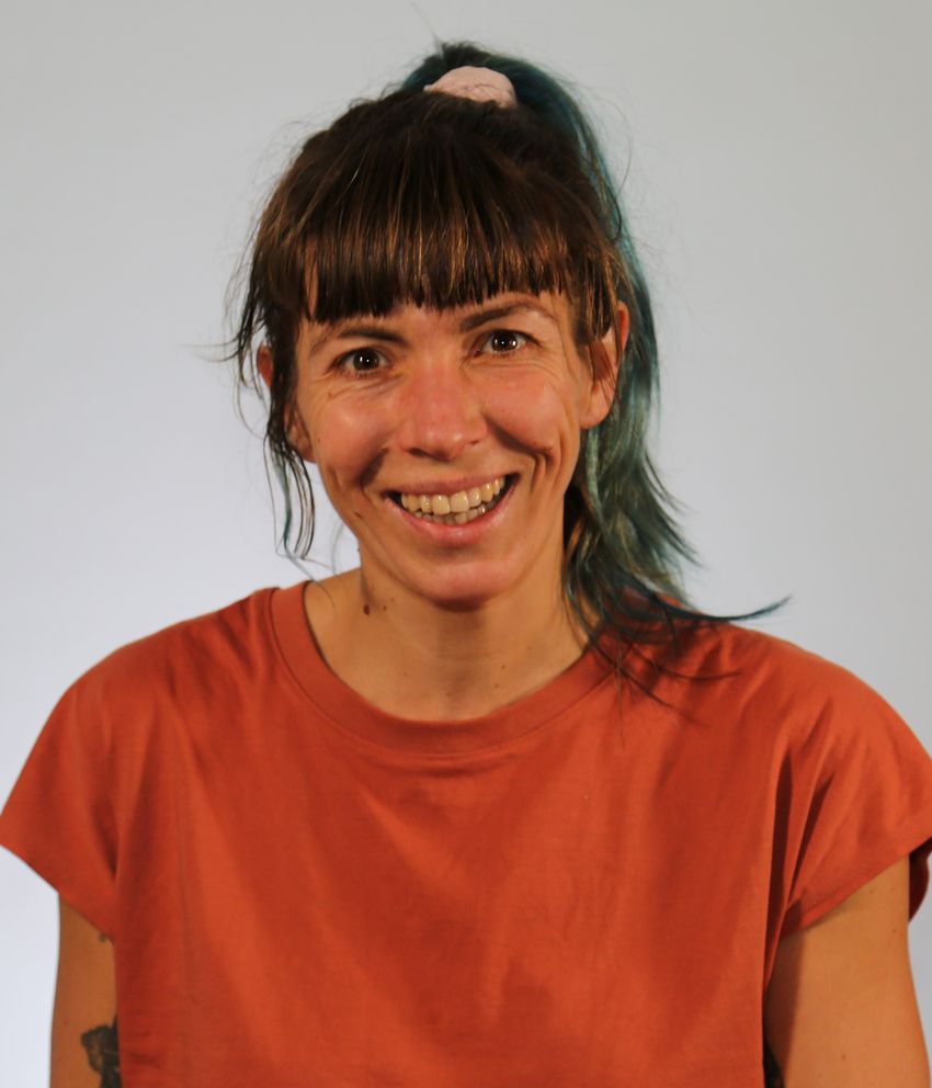 Milena Foerster, wearing an orange T-shirt, smiles at the camera. A tattoo on her right upper arm is partially visible.