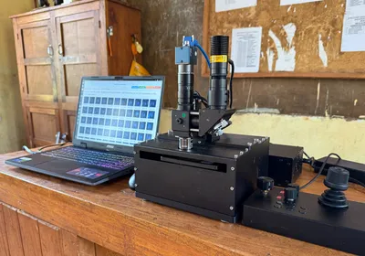 Photograph of Prakash’s low-cost, AI-powered self-driving microscope sitting on a wooden desk beside a laptop opened to show images from a slide that was recently scanned. The microscope looks like a black box with a slot near the top where a movable arm comes out to receive a glass slide. The lens extends from the top of box like a tower. On the right of the microscope, a black device with toggles and a hand held controller sits to allow workers to move the microscope around the slide and control its functions. 