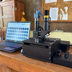 Photograph of Prakash’s low-cost, AI-powered self-driving microscope sitting on a wooden desk beside a laptop opened to show images from a slide that was recently scanned. The microscope looks like a black box with a slot near the top where a movable arm comes out to receive a glass slide. The lens extends from the top of box like a tower. On the right of the microscope, a black device with toggles and a hand held controller sits to allow workers to move the microscope around the slide and control its functions.