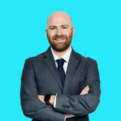 Photograph of Jevin Lortie standing in front of a bright blue background. He is wearing a dark grey suit with a white collared undershirt and dark tie. He has his arms crossed and is smiling at the camera. He also wears a watch and has a short beard. 