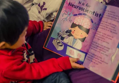 A young child holding one of the CuSTEMized books that is open to a page titled ‘N is for Neuroscientist.’
