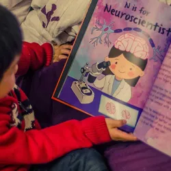 A young child holding one of the CuSTEMized books that is open to a page titled ‘N is for Neuroscientist.’