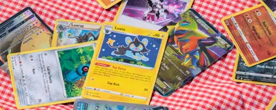 Photograph of different Pokémon cards spread disorderly out over a red and white checked blanket. 