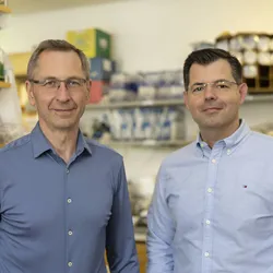 Petros Giannikopoulos (right) and Fyodor Urnov (left) stand in a lab where an on-demand therapy was made to treat Baby KJ. They are both wearing blue shirts and glasses.