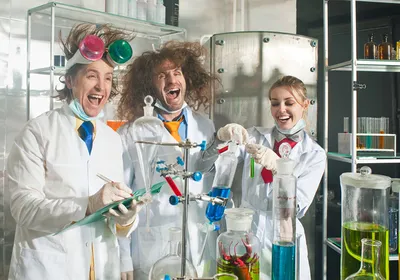A group of three cheerful chemists stand in a laboratory. They stand behind glassware filled with colorful liquids. The three scientists are laughing and the left and center figures have wild hair.
