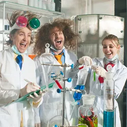 A group of three cheerful chemists stand in a laboratory. They stand behind glassware filled with colorful liquids. The three scientists are laughing and the left and center figures have wild hair.