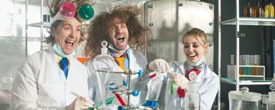 A group of three cheerful chemists stand in a laboratory. They stand behind glassware filled with colorful liquids. The three scientists are laughing and the left and center figures have wild hair.